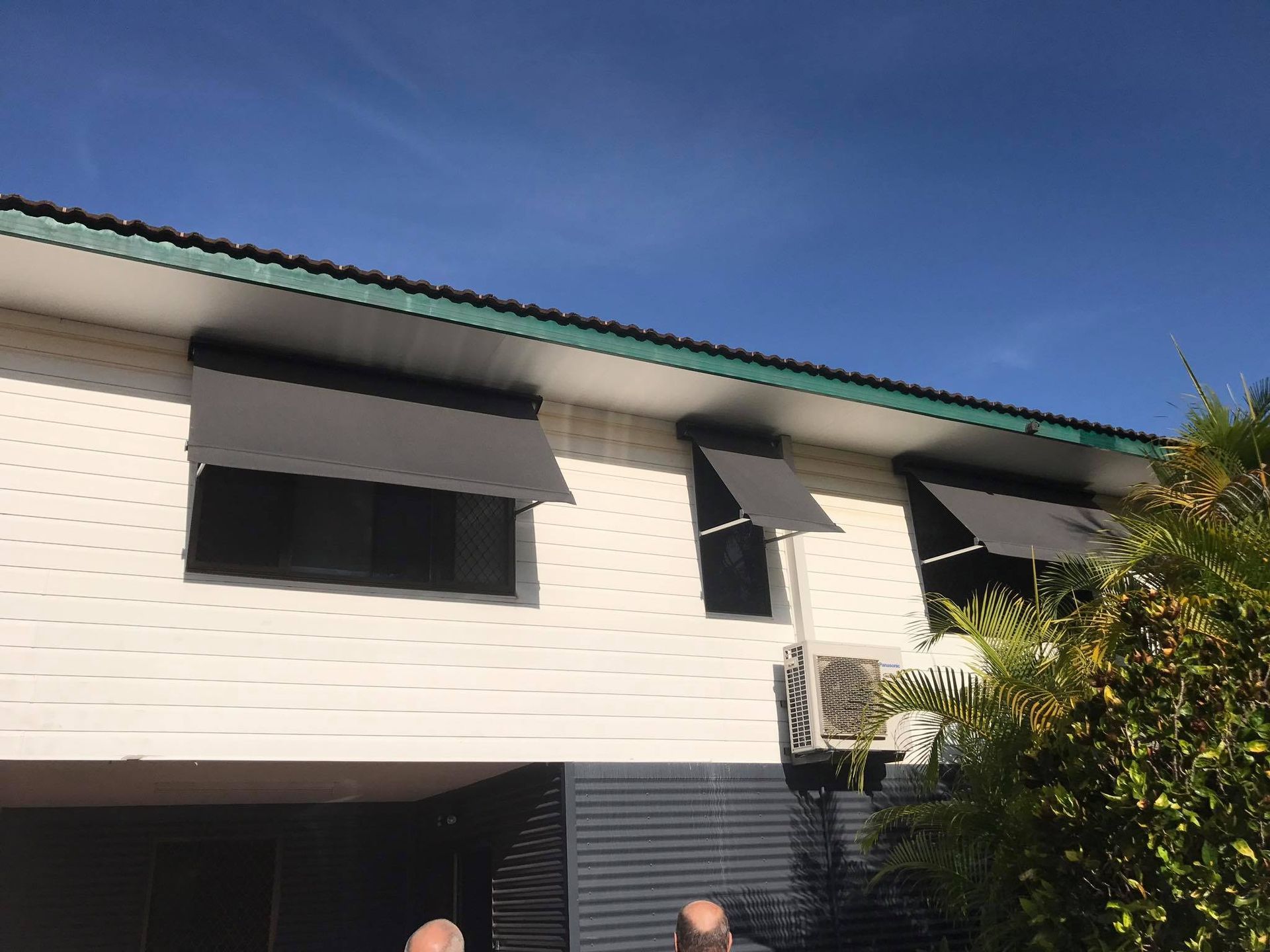 Two-story white house with black awnings and a blue sky above, partly obscured by trees and shrubs — NT Blinds in Yarrawonga, NT