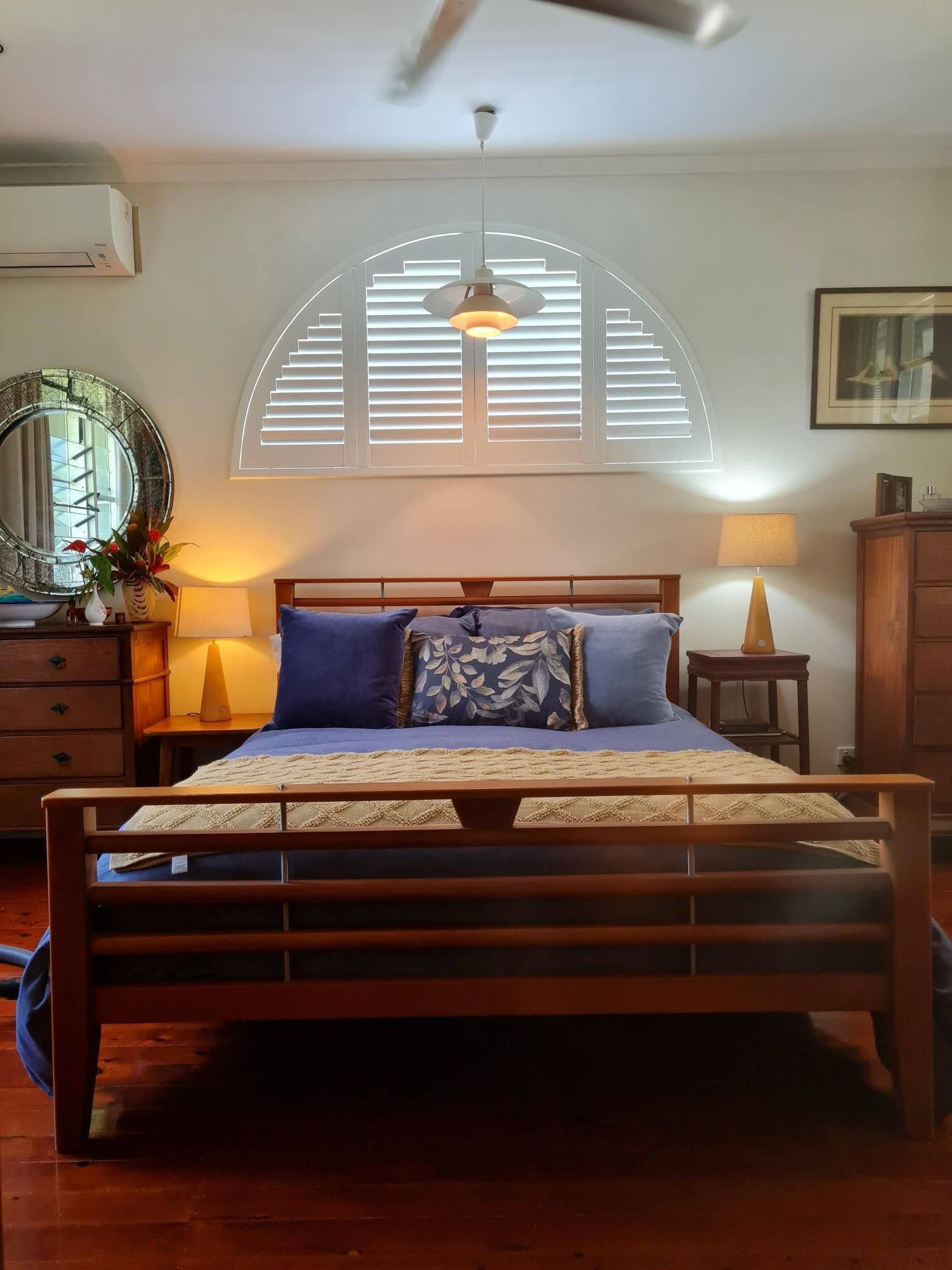Bedroom with wooden furniture, shuttered arched window, and bed with blue bedding and pillows  — NT Blinds in Palmerston, NT