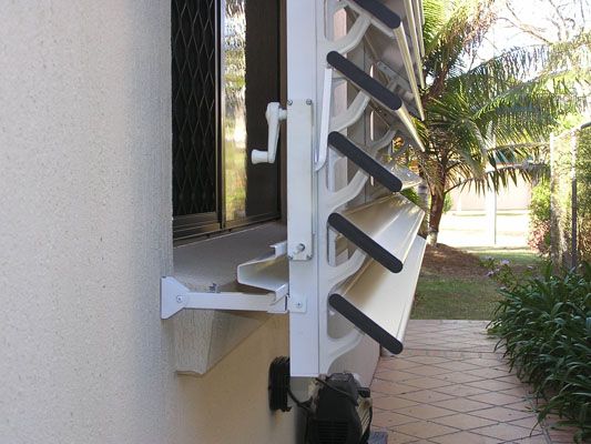 White louvered window open on a building's exterior. A hand crank controls the louvers, with a garden visible behind.