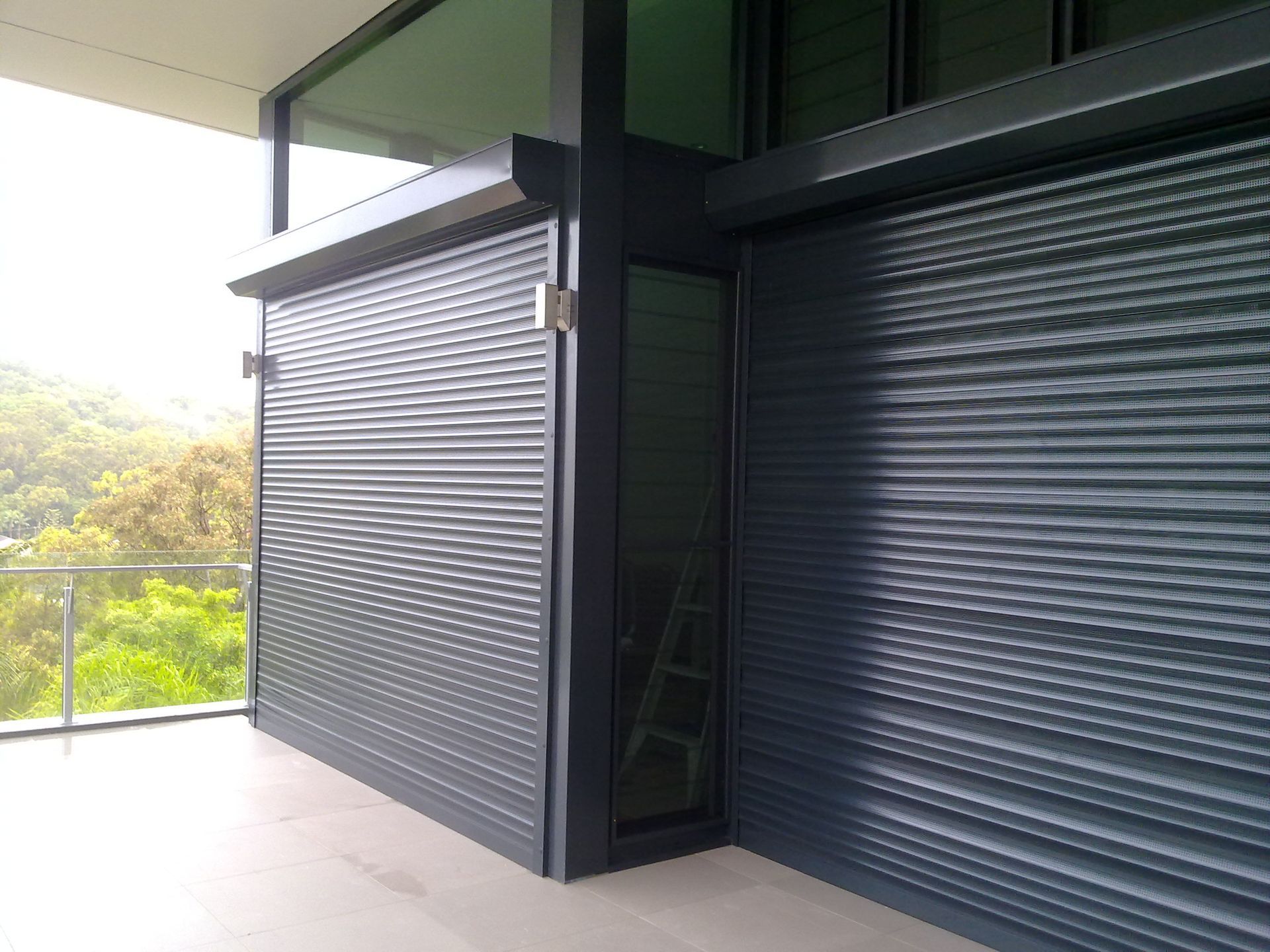 Exterior view of a building with dark gray, closed rolling shutters covering windows, and a glass balcony railing  — NT Blinds in Yarrawonga, NT