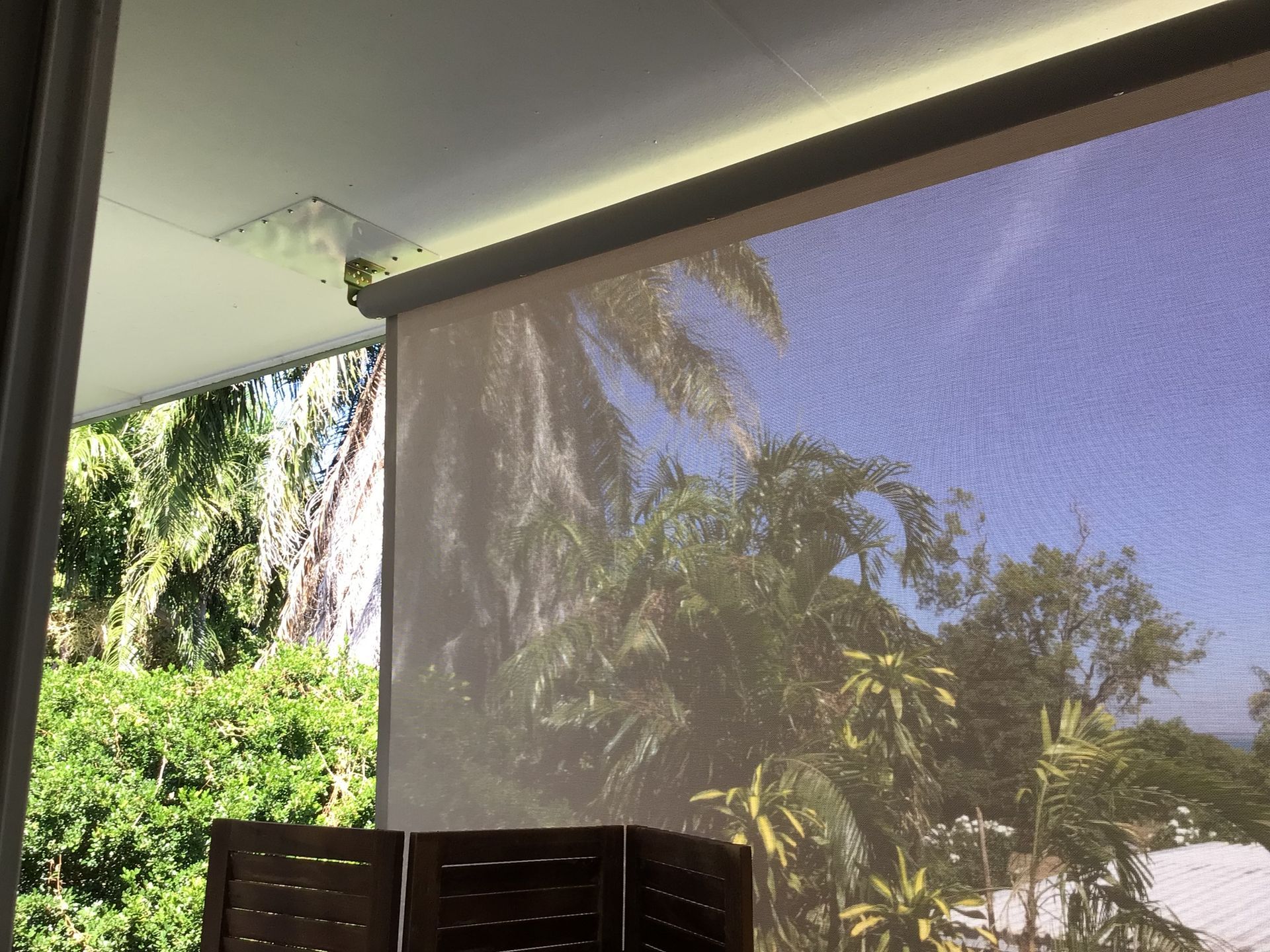Roller shade over a porch, with a view of trees and a blue sky; brown wooden screen in the foreground  — NT Blinds in Casuarina, NT