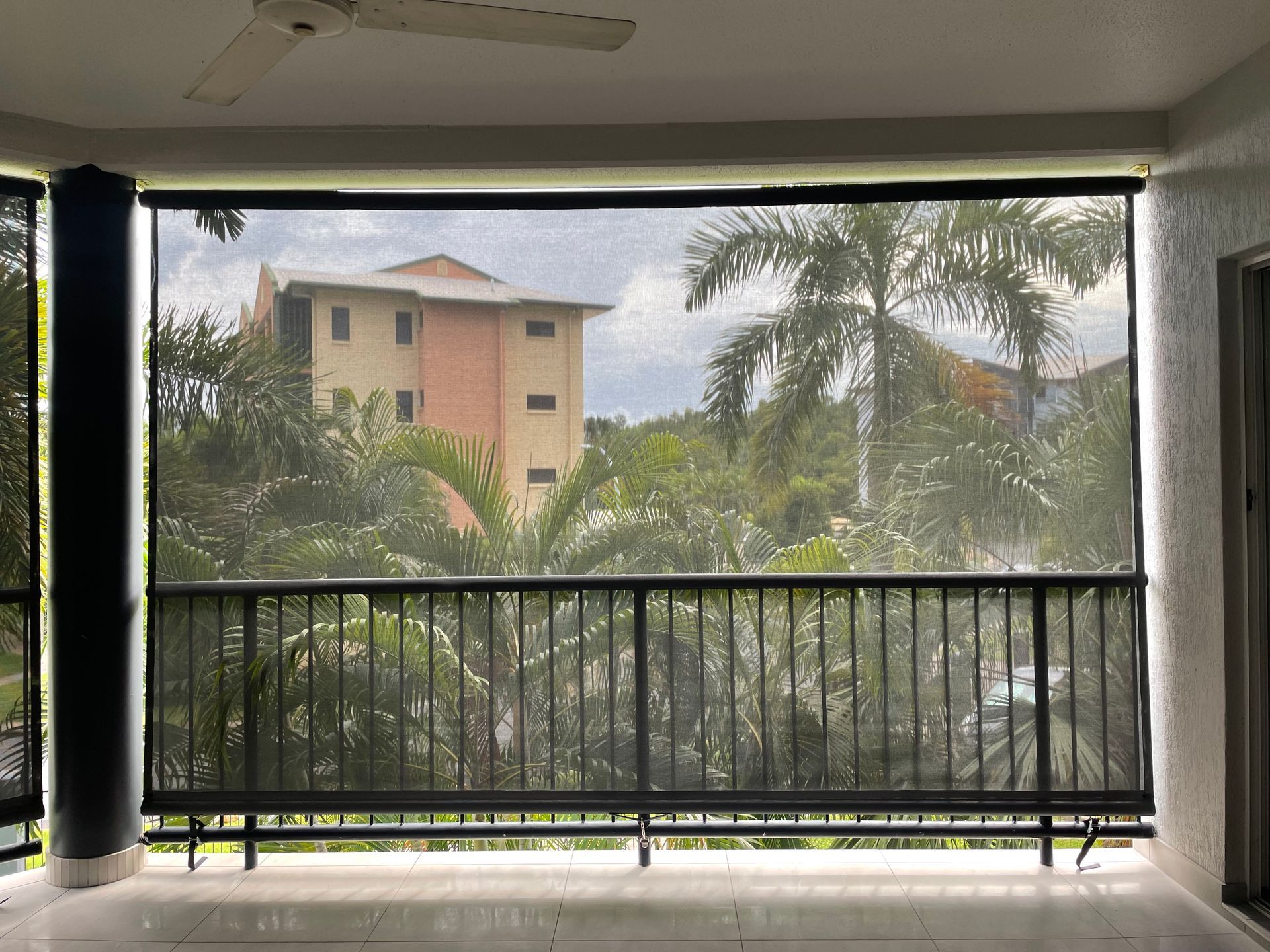 Balcony with black mesh roller shade, looking out at trees, building, and railings  — NT Blinds in Yarrawonga, NT