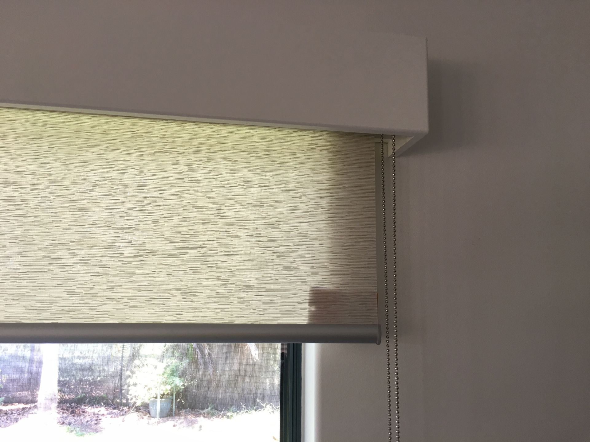 Beige roller blind partially covering a window. The blind has a chain to raise and lower it.