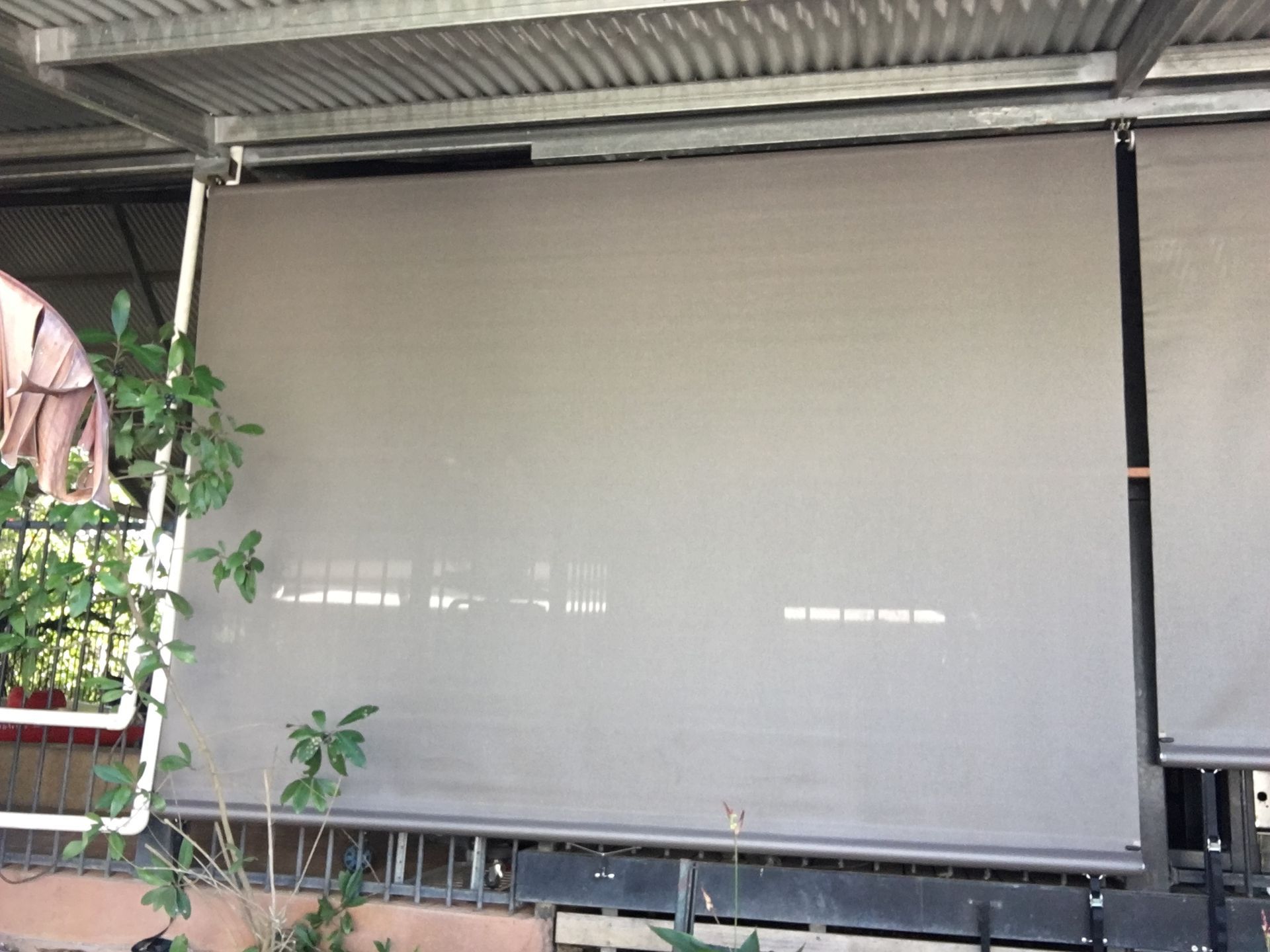 Outdoor roller shades, beige, partially lowered on a patio under a corrugated metal roof  — NT Blinds in Humpty Doo, NT