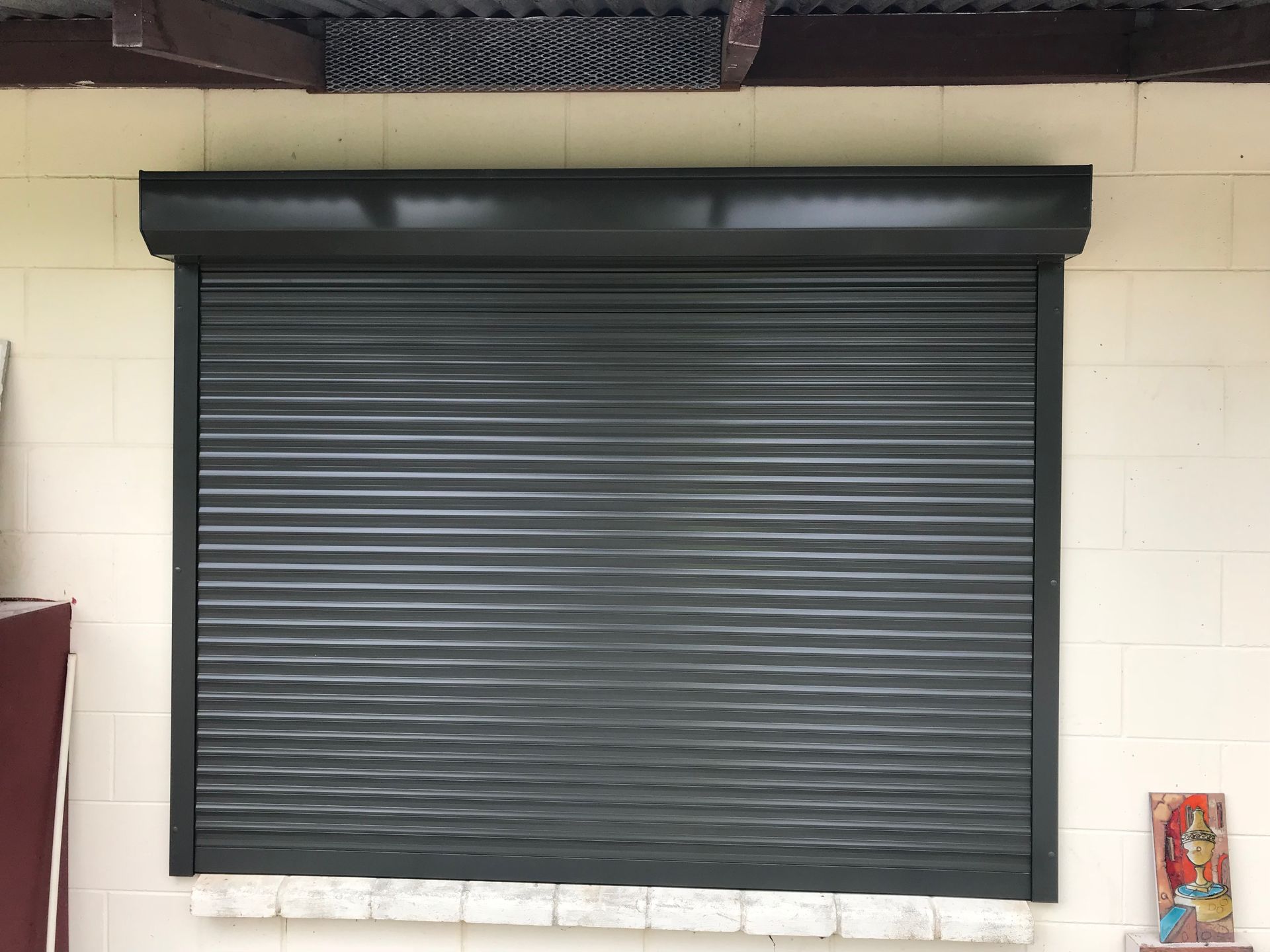 Black roller shutter over a window on a light brick wall  — NT Blinds in Yarrawonga, NT