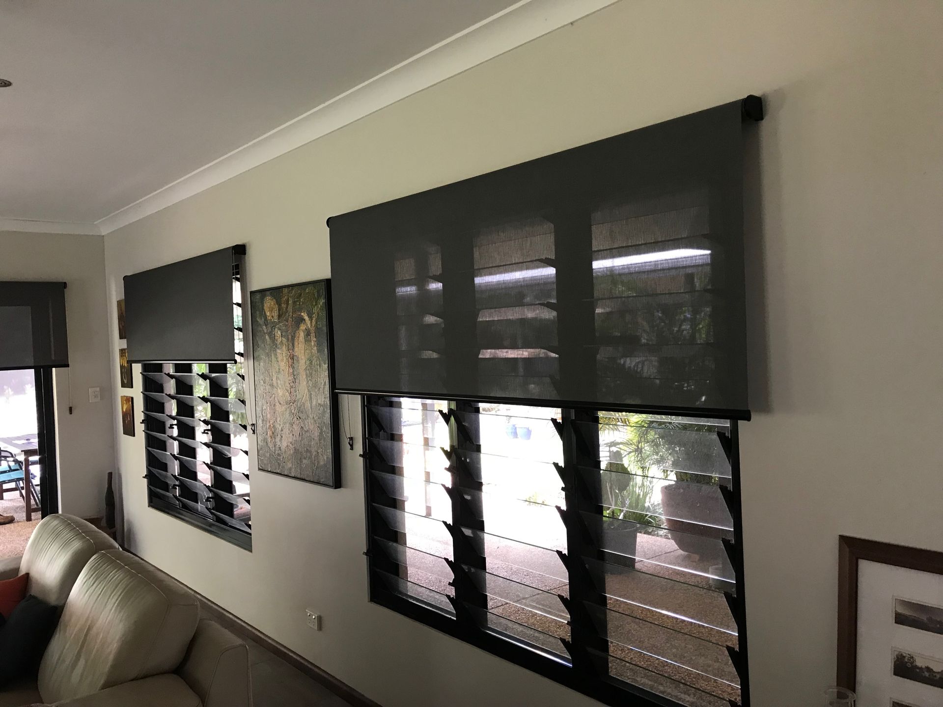 Dark grey roller blinds installed above glass louvre windows on a beige wall — NT Blinds in Fannie Bay, NT