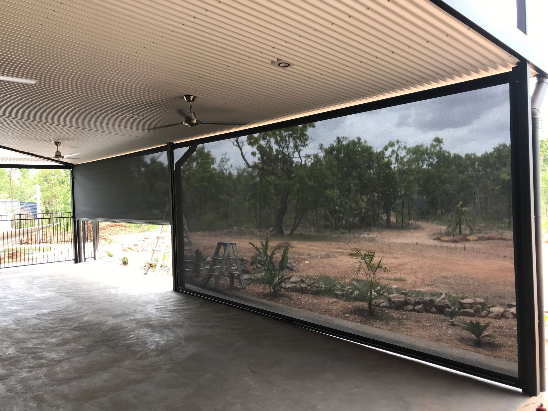 Outdoor patio with a retractable black screen, concrete floor, and ceiling fan; view of trees  — NT Blinds in Fannie Bay, NT