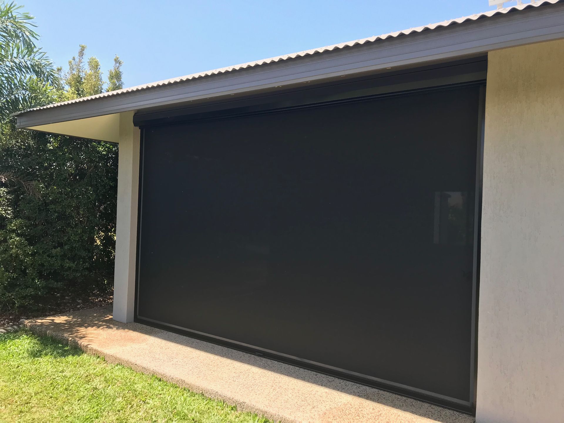 Black outdoor roller shade on a white building, partially covering a doorway. Green grass and blue sky are visible — NT Blinds in Howard Springs, NT