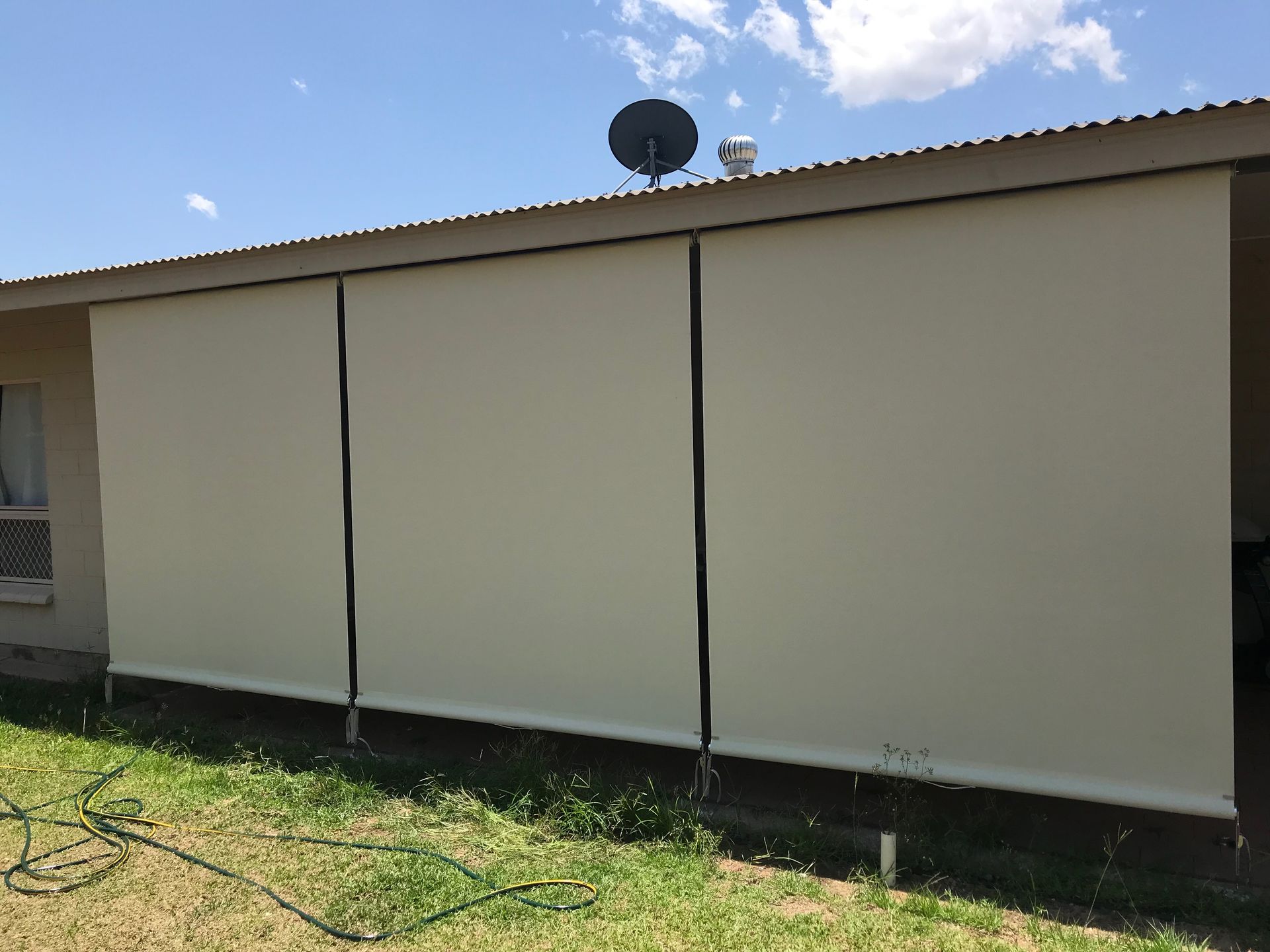 Three beige outdoor roller shades installed on the side of a building, with a grassy lawn in the foreground  — NT Blinds in Coolalinga, NT