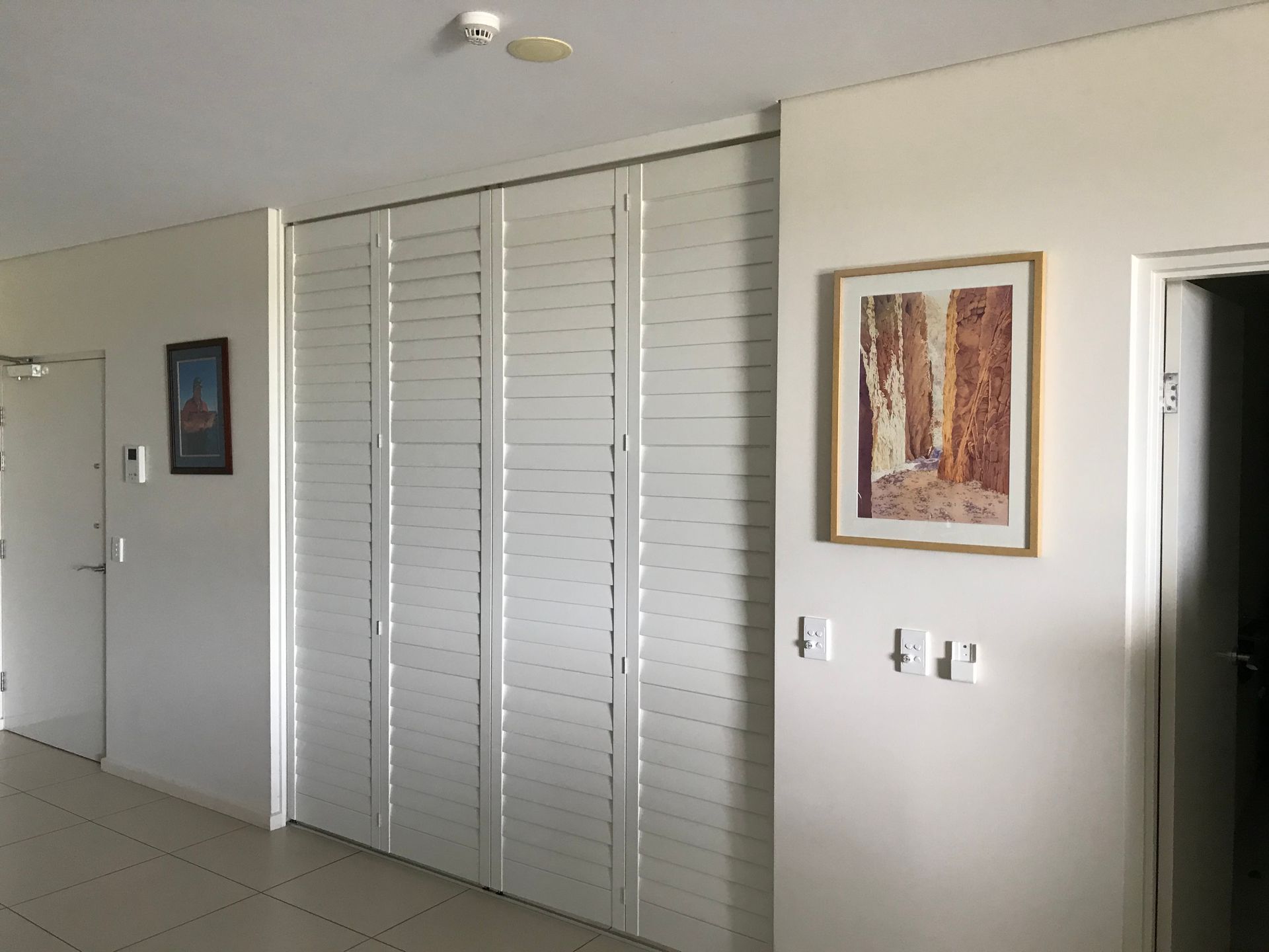 Hallway with white louvered doors, art, and an open doorway.  White walls and light-colored tiled floor.