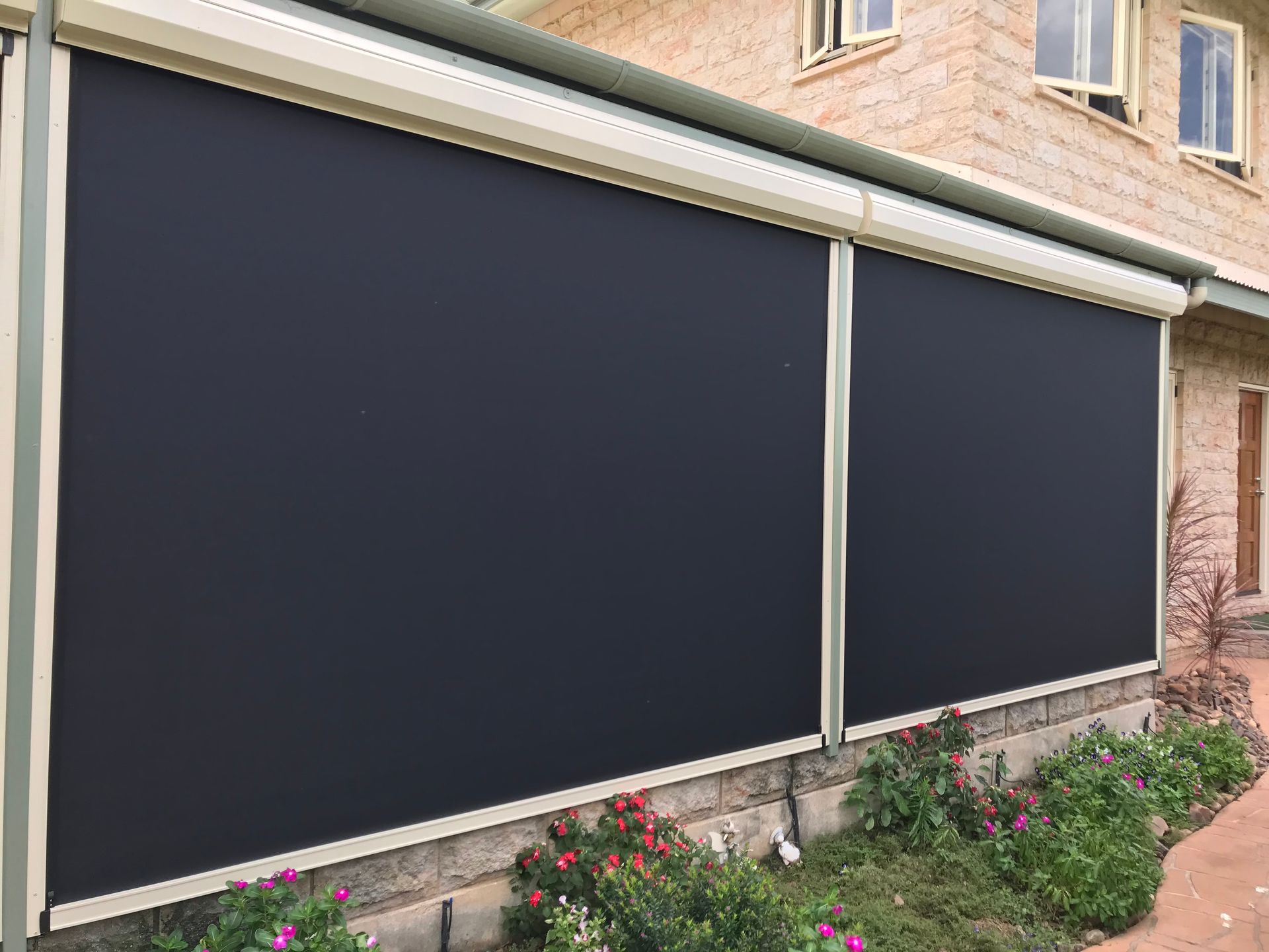 Black outdoor roller blinds covering a patio, near a brick house and garden  — NT Blinds in Darwin City, NT