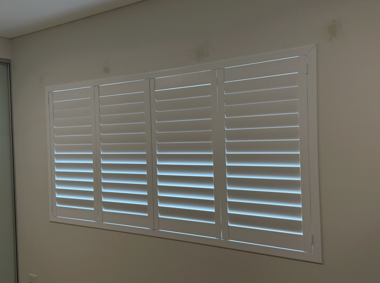 White plantation shutters on a window, partially open, letting in filtered light. Interior setting  — NT Blinds in Yarrawonga, NT