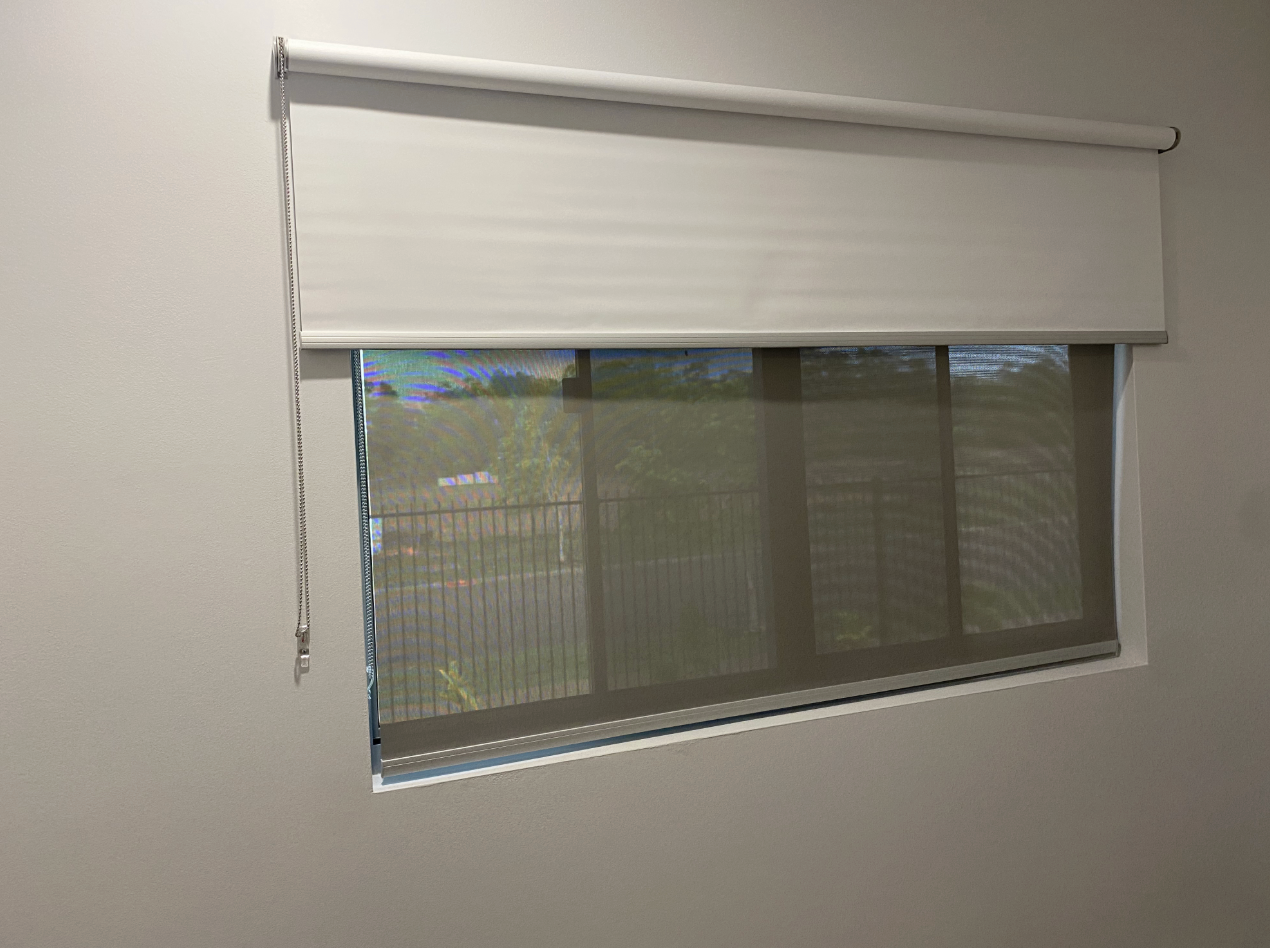 A window with dual roller shades, one open, revealing a view through the window  — NT Blinds in Howard Springs, NT