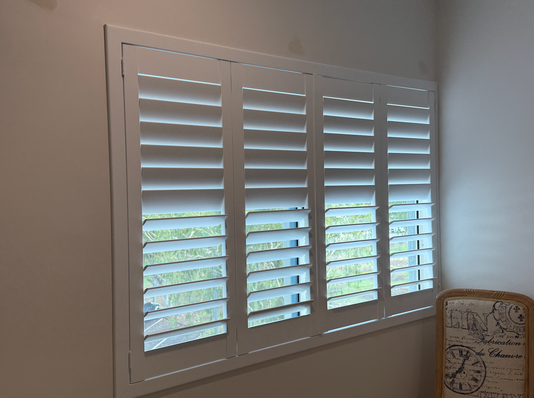 White window shutters partially closed, set in a white frame, with a glimpse of greenery outside  — NT Blinds in Yarrawonga, NT