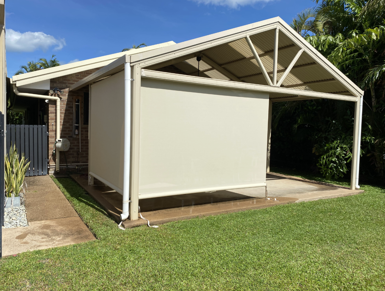 Beige awning covering a patio area with a rolled-down shade. Green lawn and brick building nearby  — NT Blinds in Yarrawonga, NT