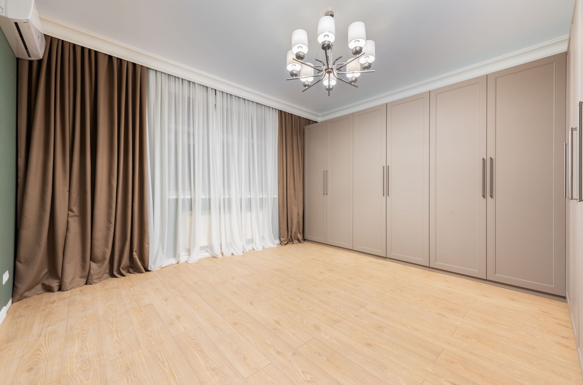 Empty beige room with wood floor, large curtains, built-in wardrobes, and a chandelier 