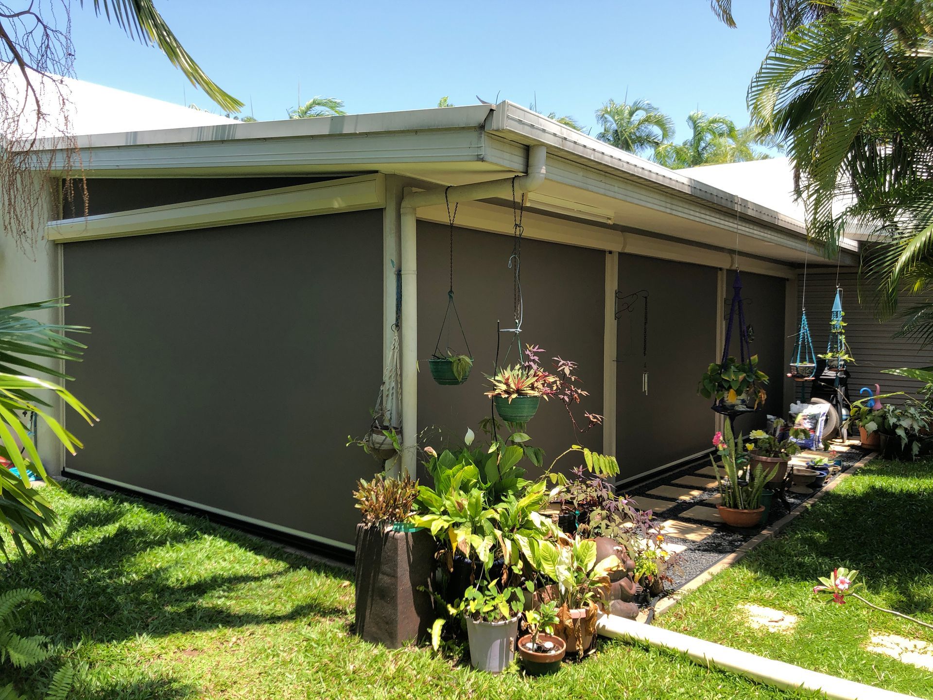 Brown outdoor shades enclose a patio, surrounded by plants and green grass  — NT Blinds in Yarrawonga, NT