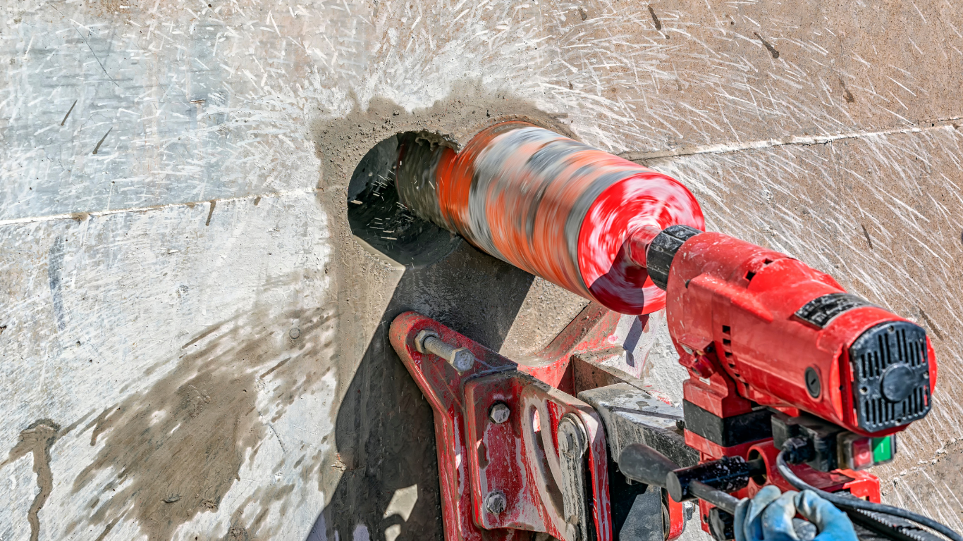 Red power drill cutting a circular hole in a concrete wall with dust and debris nearby