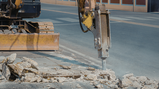 Construction excavator jackhammering concrete on a road, breaking up pavement near a curb