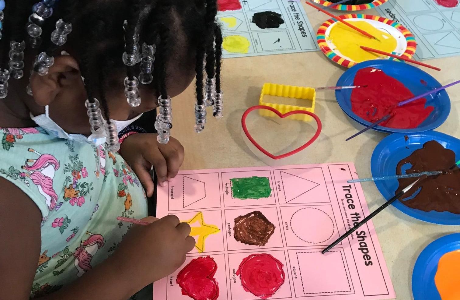 A little girl is painting on a piece of paper that says trace the shapes