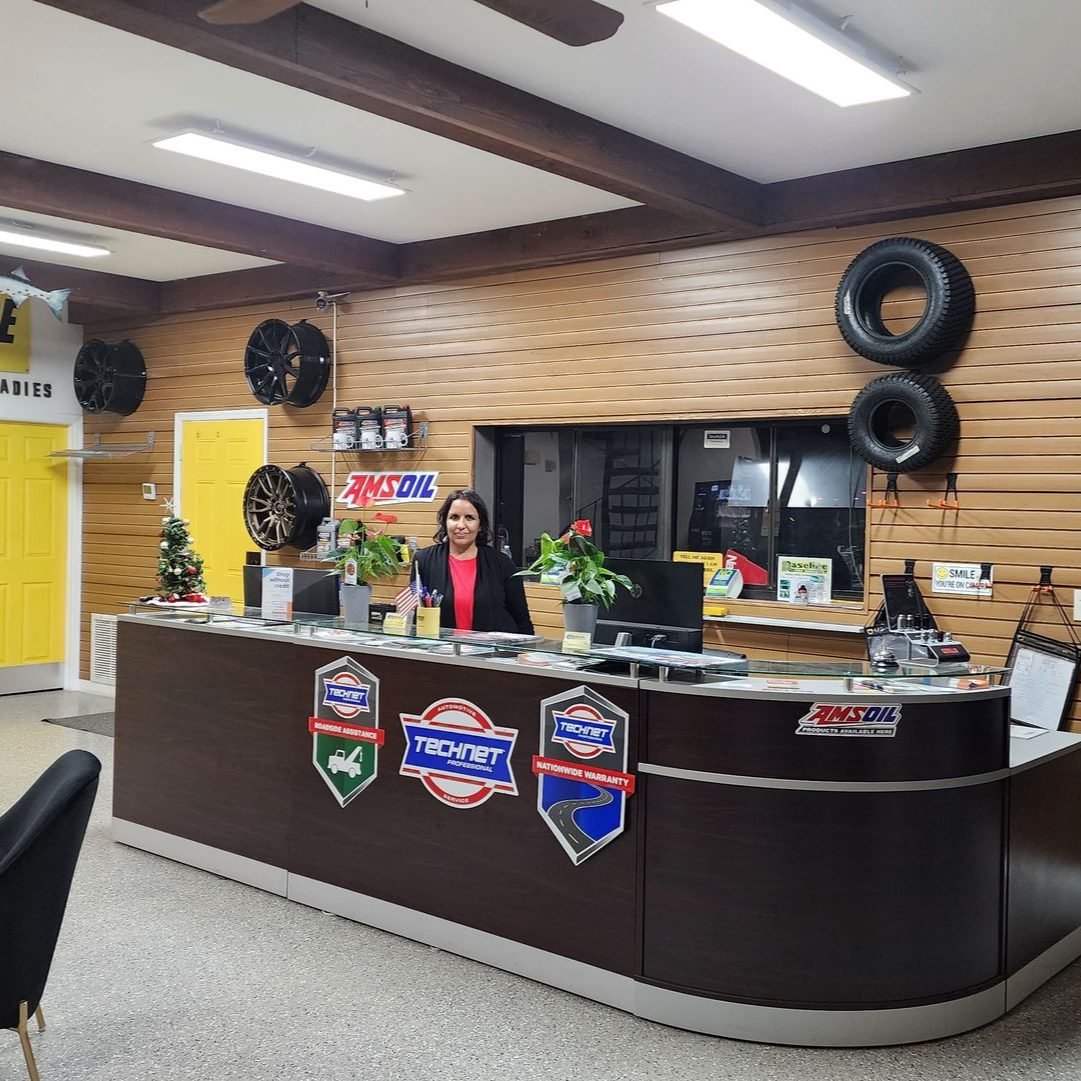 Woman at auto shop counter, brown and beige tones, AMSOIL logos, tires displayed, small Christmas tree.