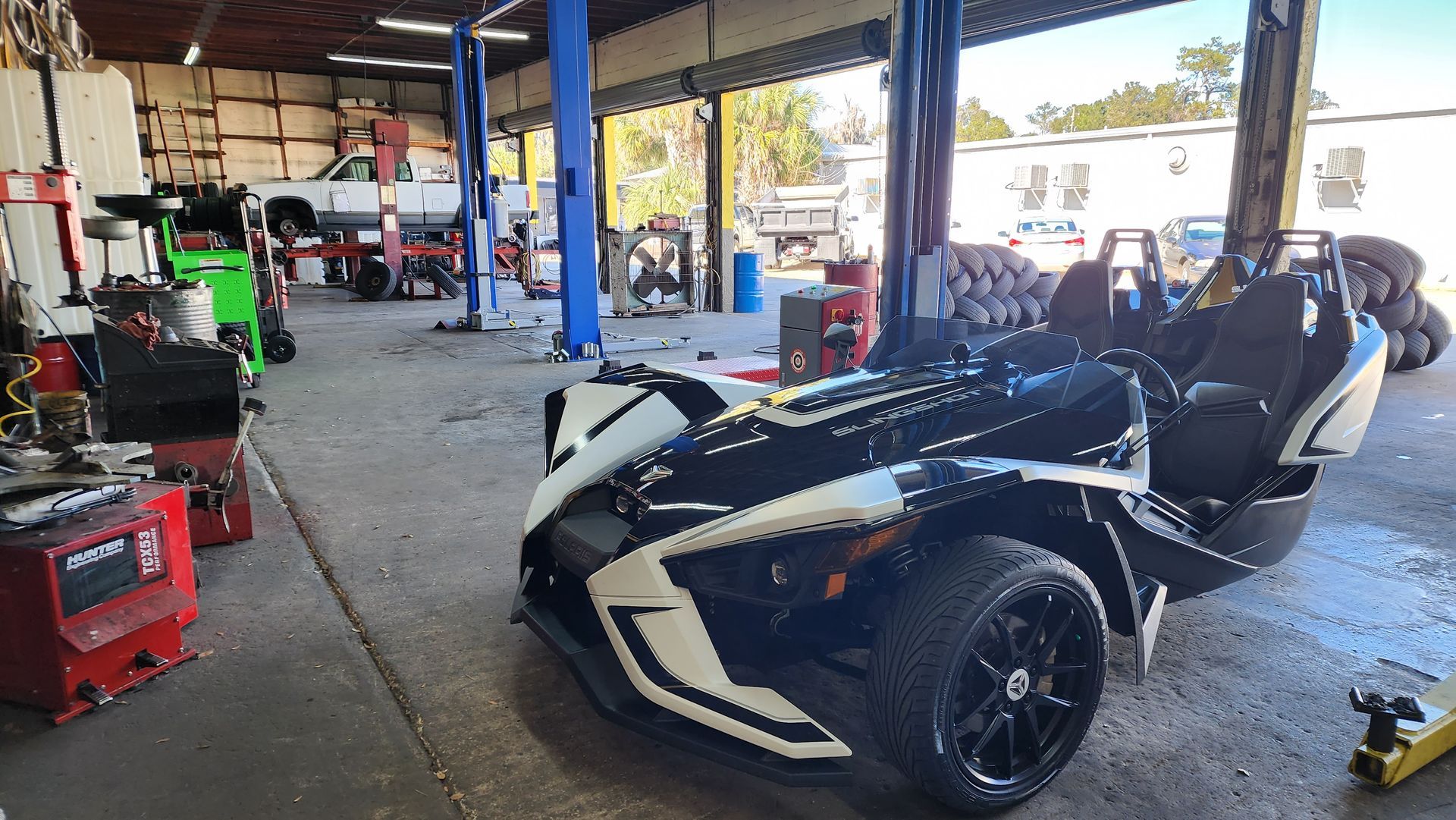 A black and white Polaris Slingshot in a garage. Shop equipment and lifted vehicle in the background.