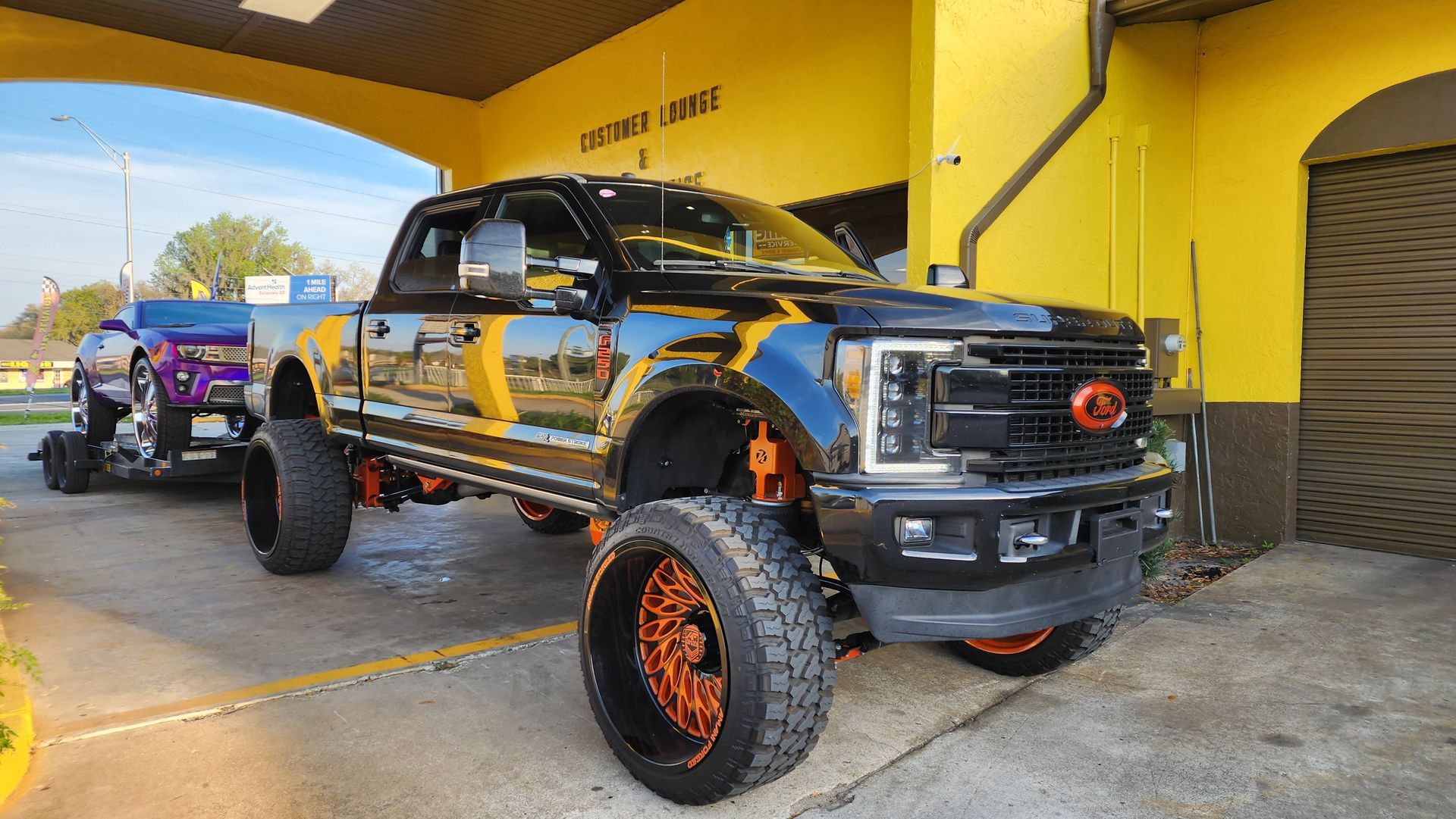 Black and orange lifted truck parked under yellow archway, towing a purple car.