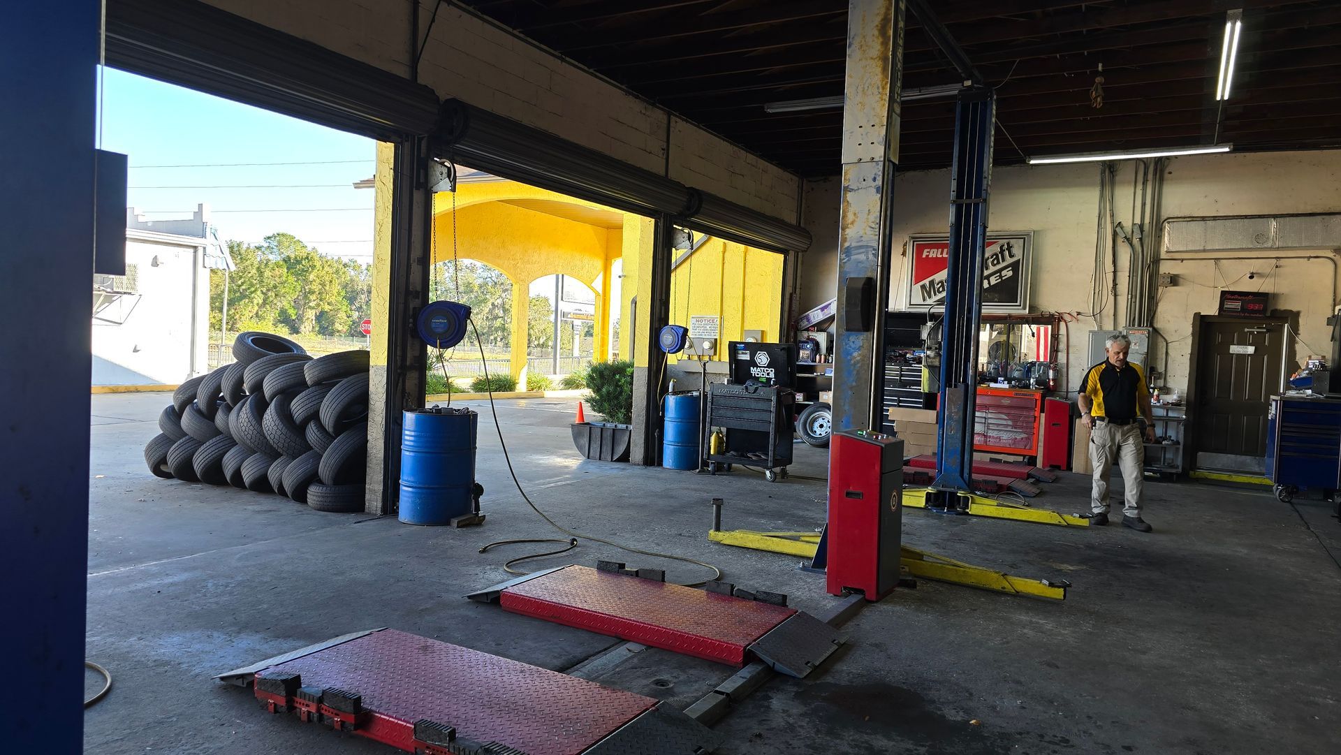 Inside a repair shop with open doors; tires, tools, and a car lift are visible. A person stands nearby.