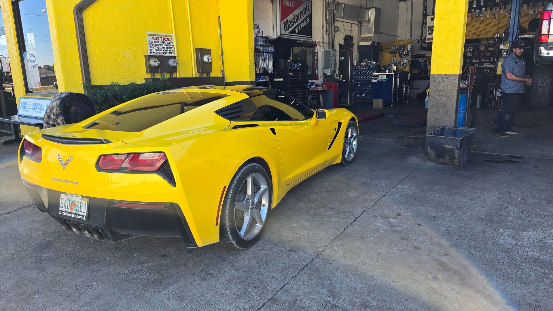 Yellow Corvette sports car in a garage bay, next to an open garage door; mechanics visible.