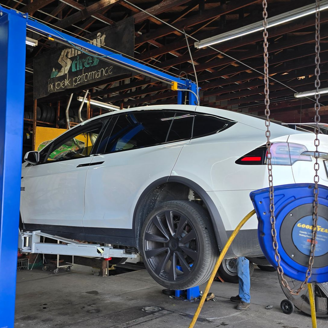 White Tesla SUV on a lift in a repair shop, with blue lift arms and a cable reel.