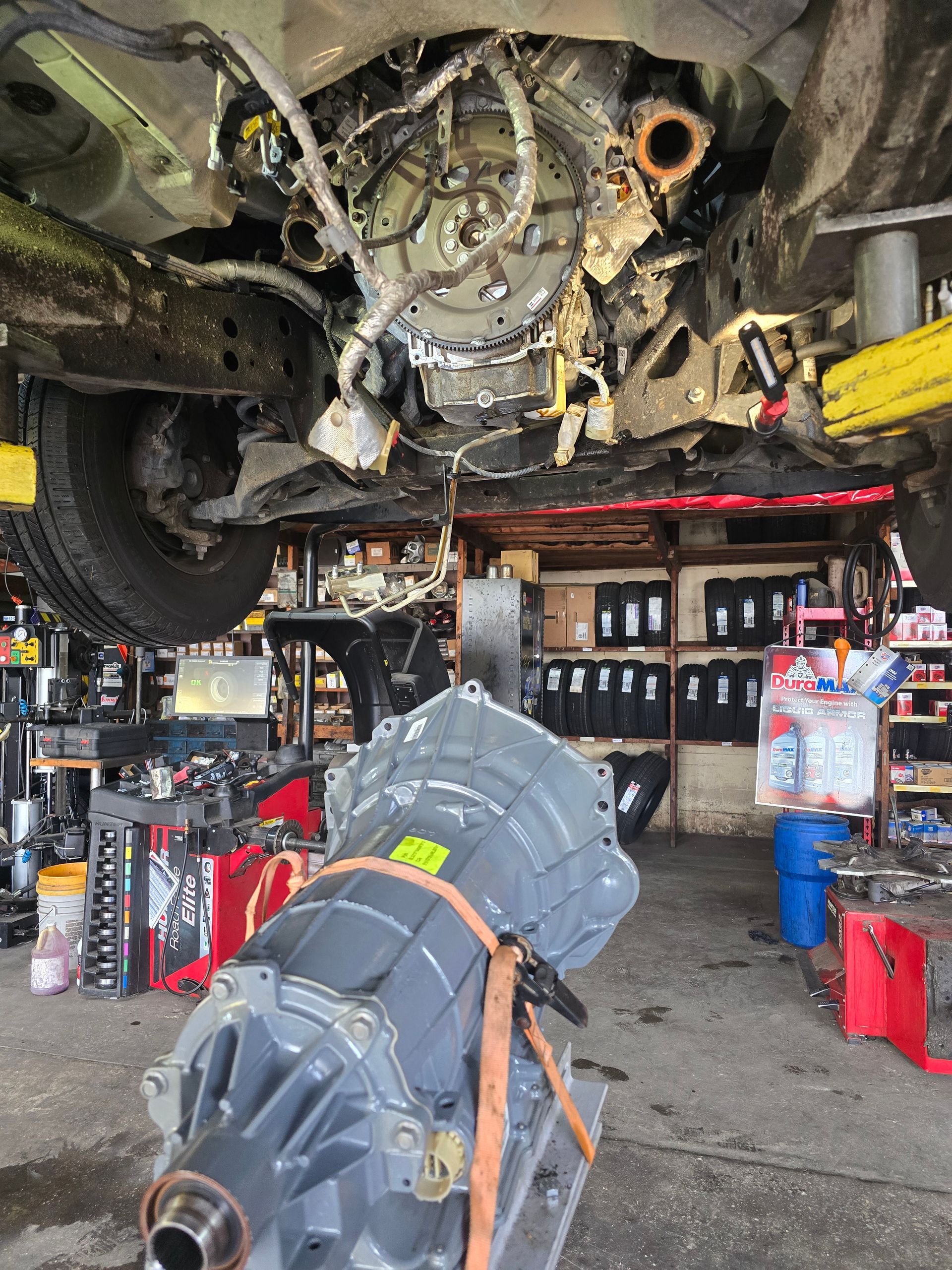 A car is elevated on a lift in a repair shop. A new transmission sits on the floor.