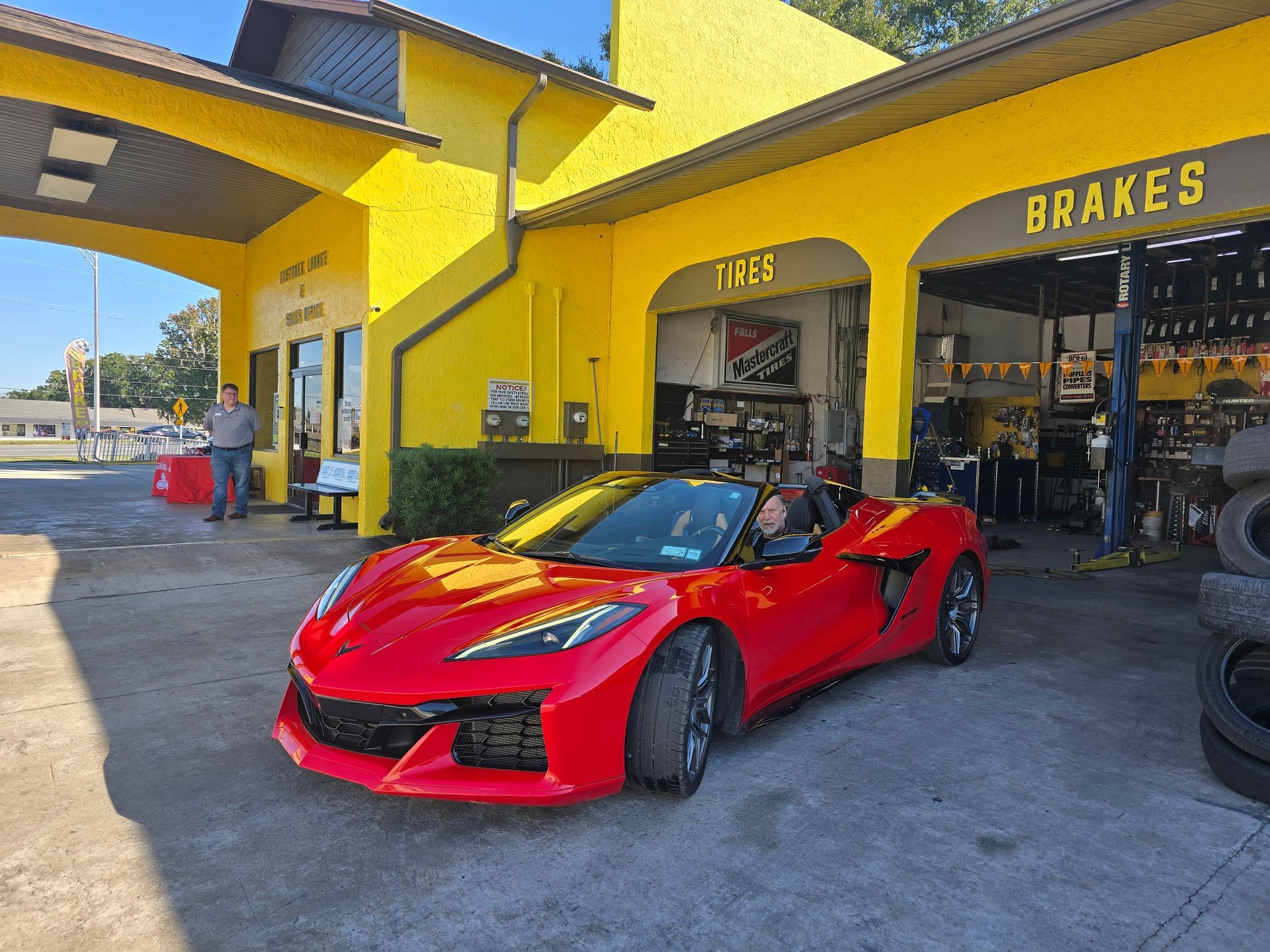 Red Corvette at a yellow auto repair shop; driver inside.