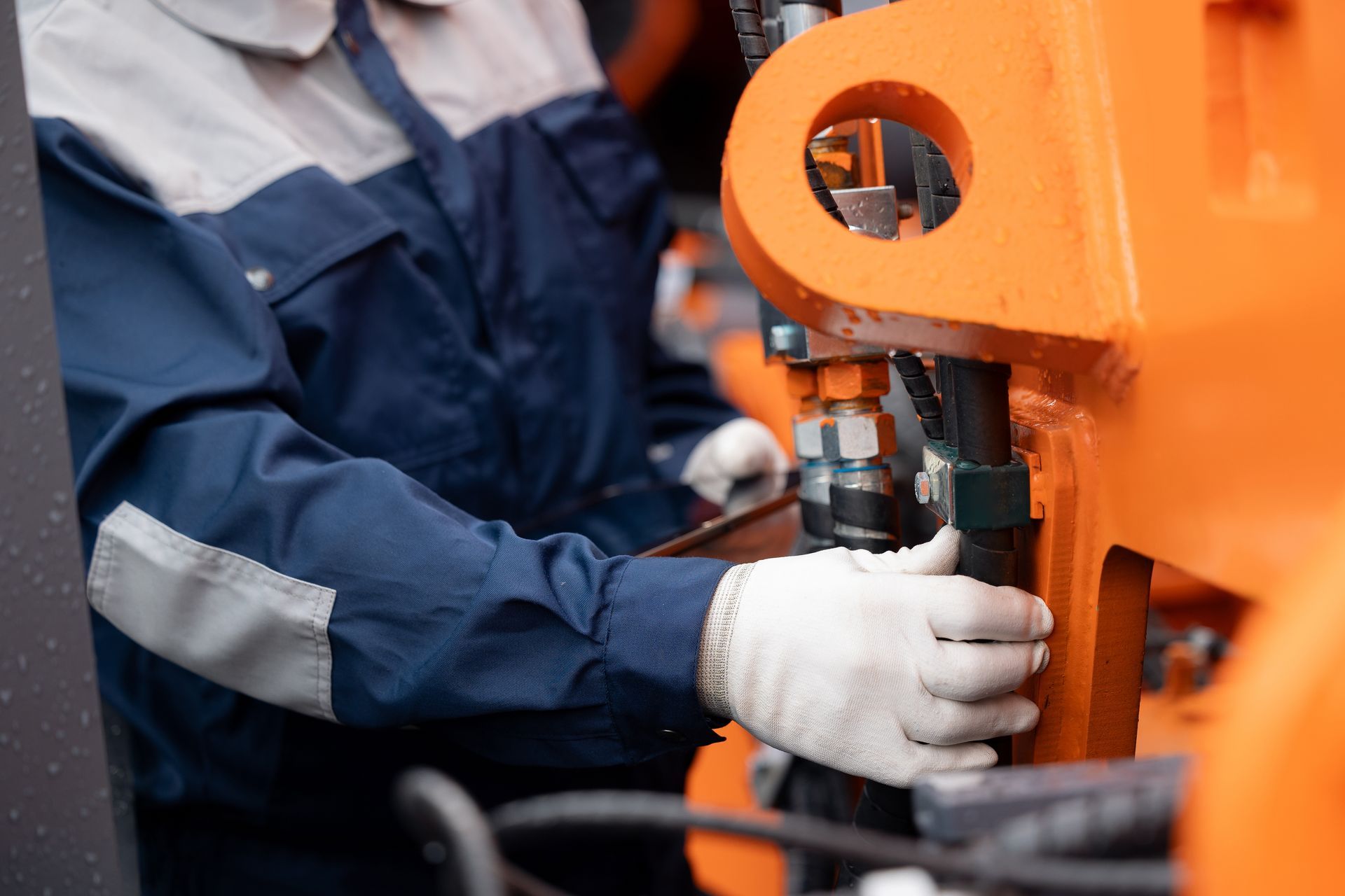 Mechanic in blue uniform, white gloves, repairing orange machinery.