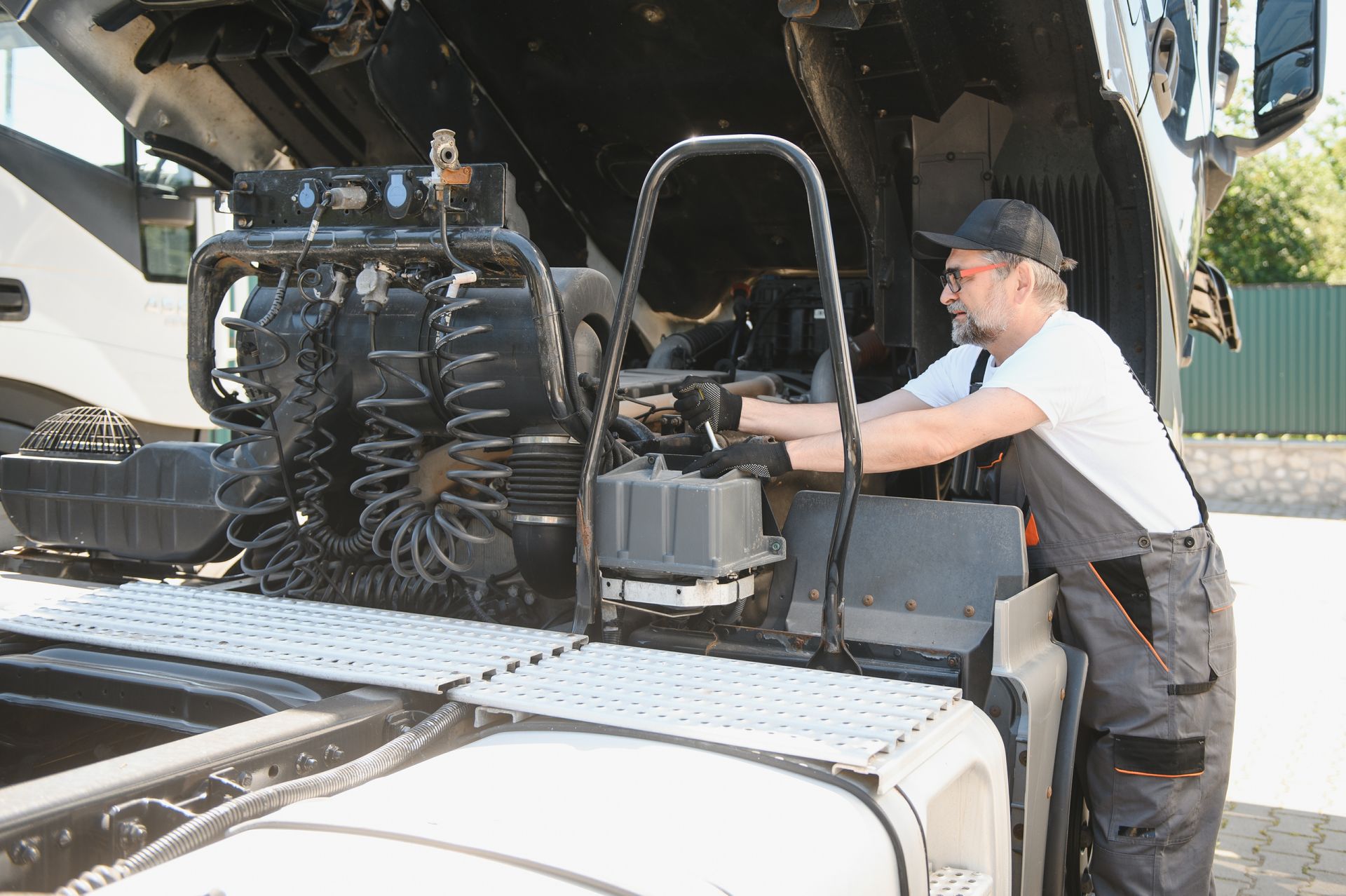 Mechanic working on the engine of a truck outdoors. He wears overalls and a cap, looking at engine components.