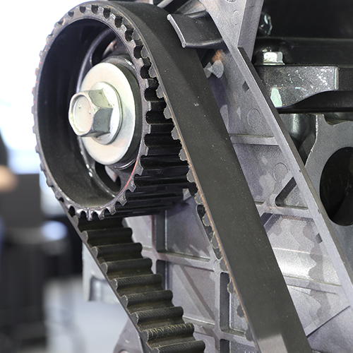 Black timing belt and pulley on an engine, visible gears and silver metal components.