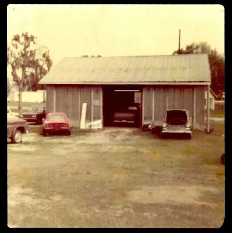 Old barn with open door, several cars parked in front, grass in the foreground.