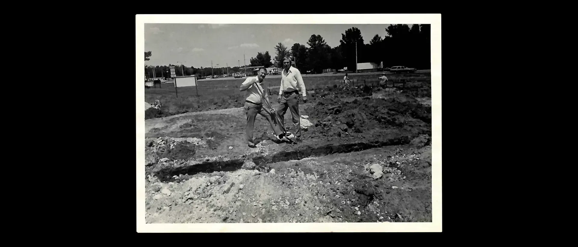 Two men standing in a dirt area, digging with a shovel, trees in the background.