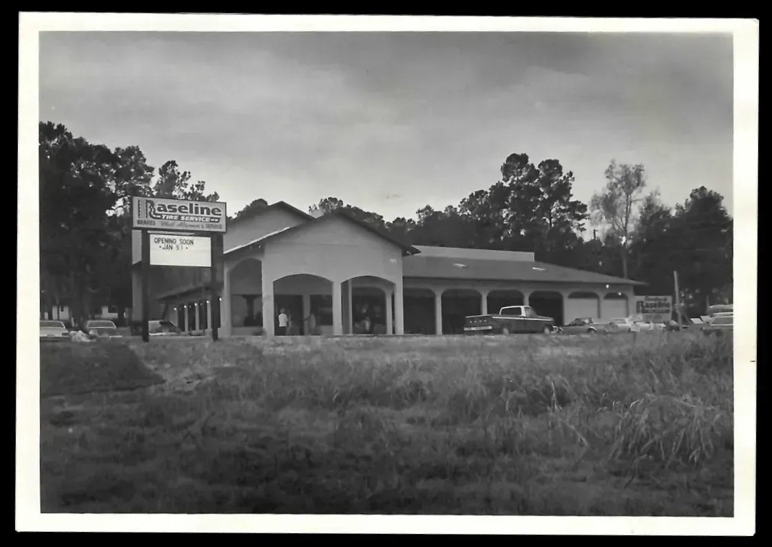 Restaurant with a sign, visible columns, and a car in front. Grassy field in the foreground, trees in the background.