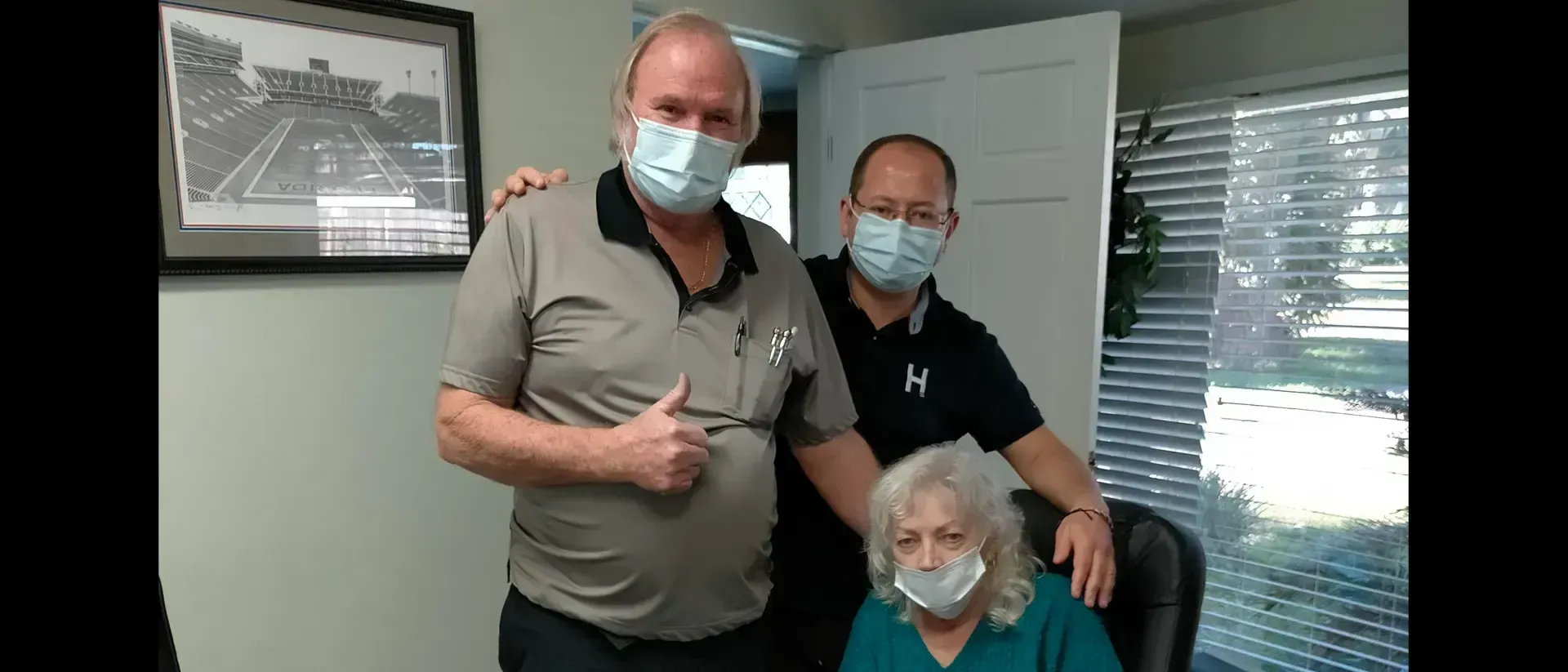 Three people in masks pose for a photo in an office. One man gives a thumbs up. A window is in the background.
