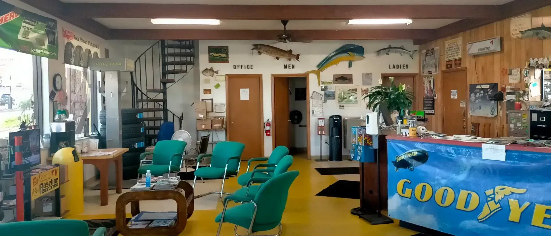 Interior of a waiting room with teal chairs, a yellow floor, and a Goodyear banner. A staircase is visible.