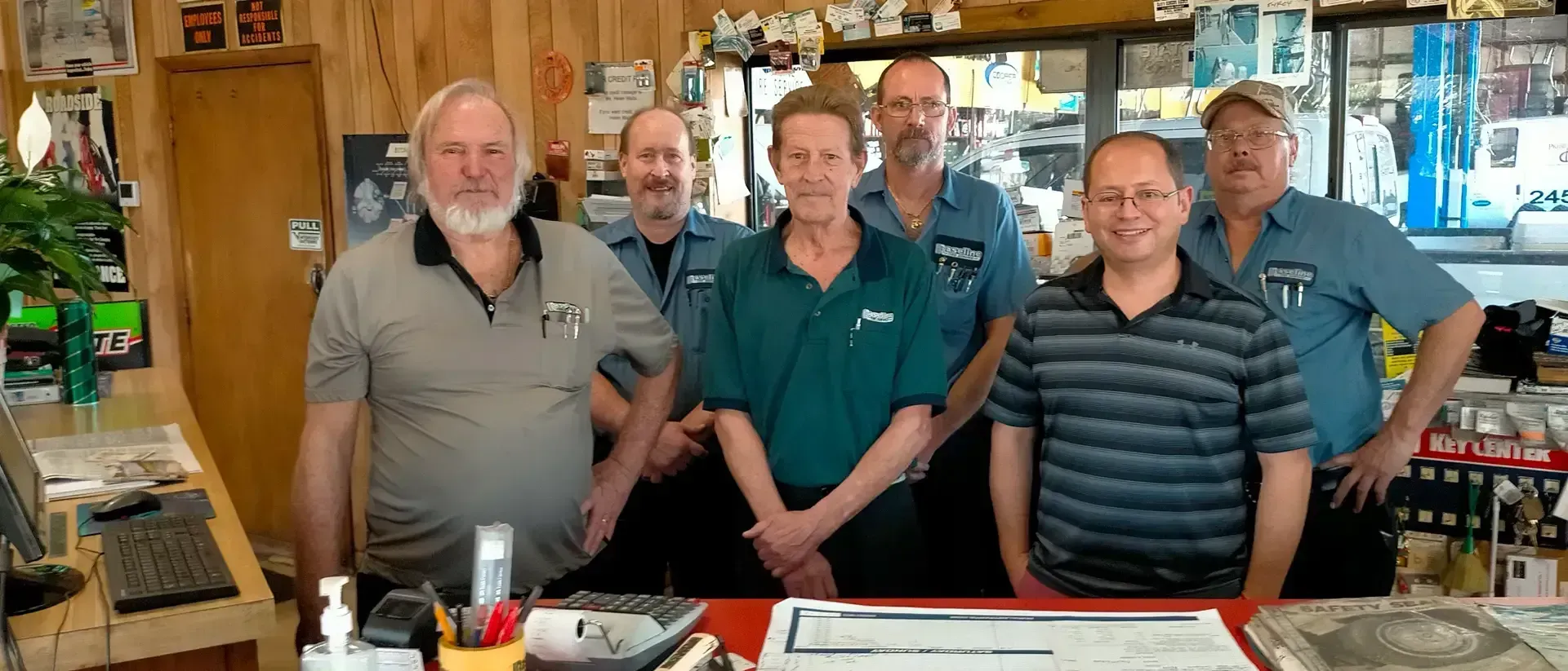 A group of six men standing behind a counter in an auto parts store. They are smiling.