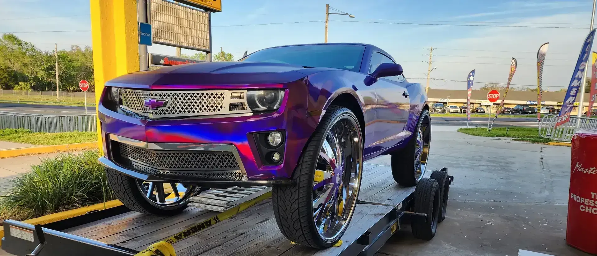 Purple Chevrolet Camaro on a trailer, with large chrome wheels, in a parking lot.