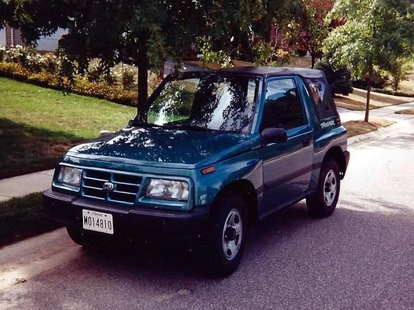 Teal two-door Geo Tracker parked on a paved street. Front view, black trim, parked near a grassy area and trees.