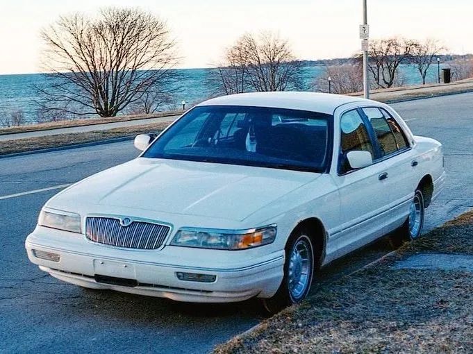 White Mercury Grand Marquis sedan parked on a roadside by a body of water.
