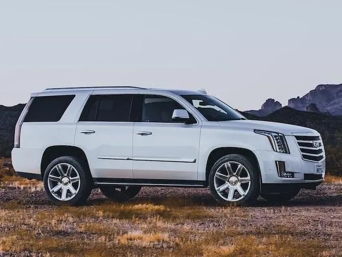 White Cadillac SUV parked on dirt road in front of hills.