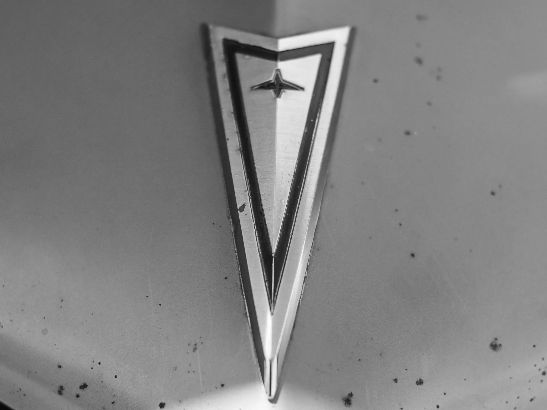 Black and white close-up of a vintage Pontiac arrowhead emblem on a car hood.