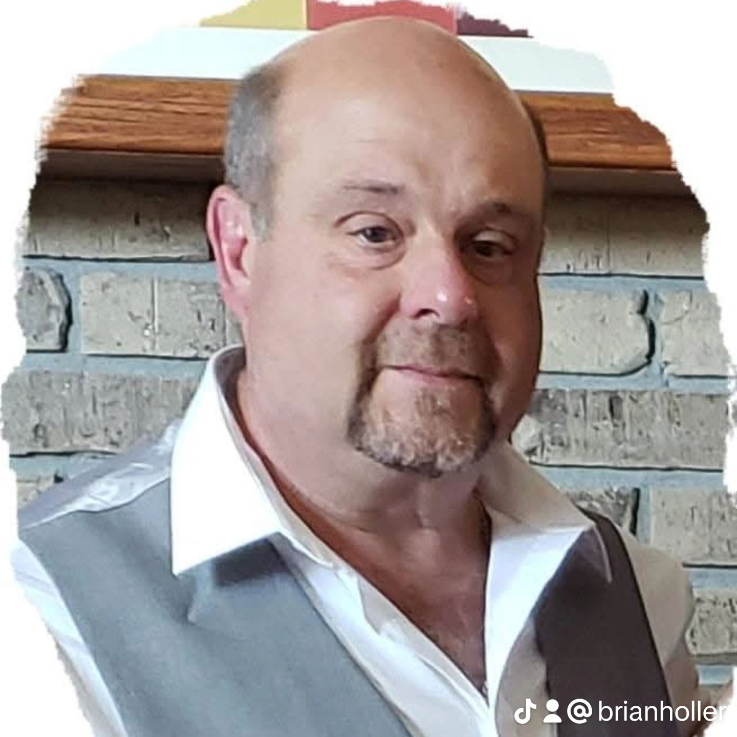 Bald man with goatee wearing a grey vest and white shirt, smiling. Brick wall background.
