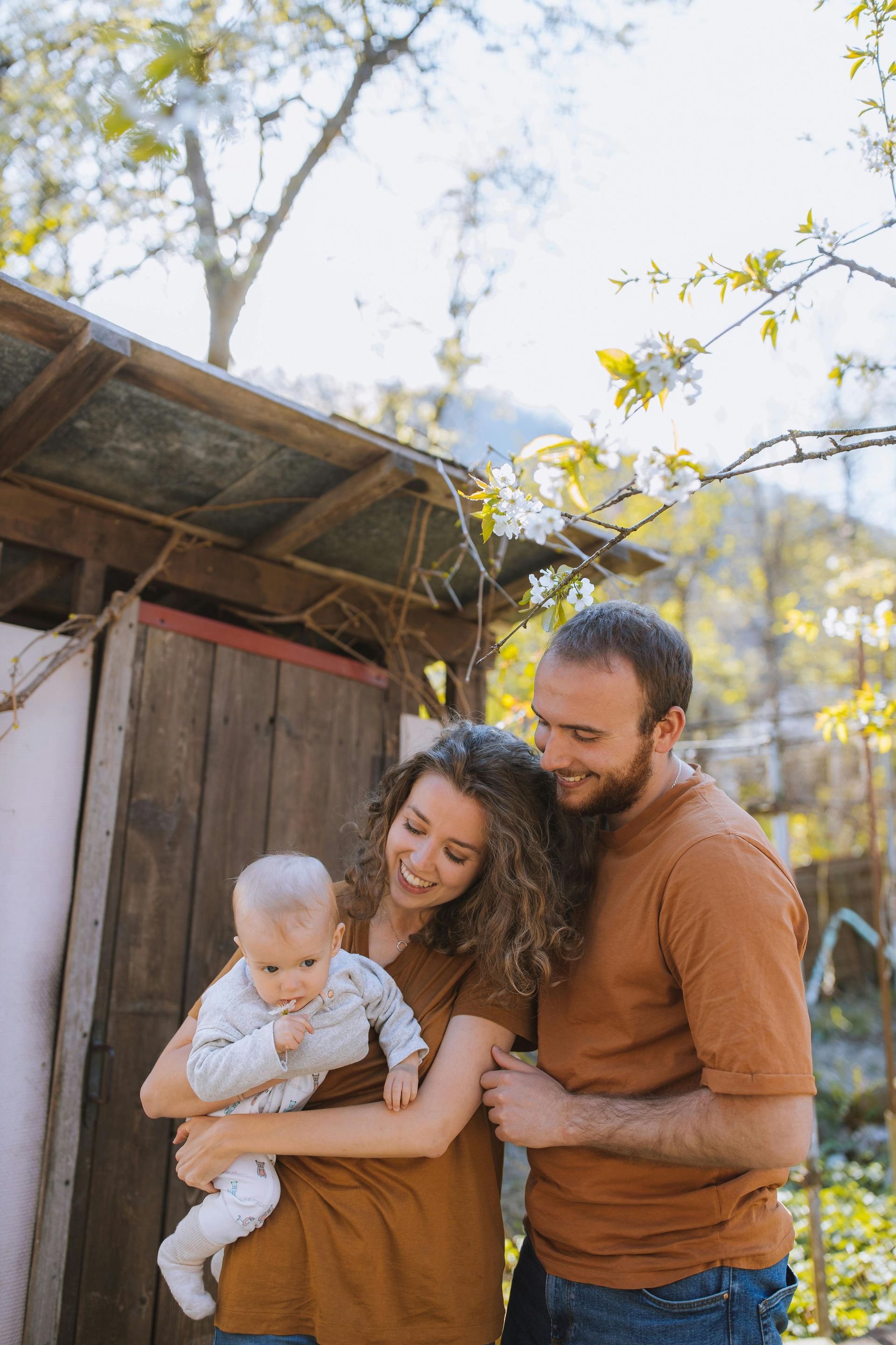 Family of three smiles outdoors. Mother holds baby, father embraces mother. Warm tones, wooden shed backdrop.
