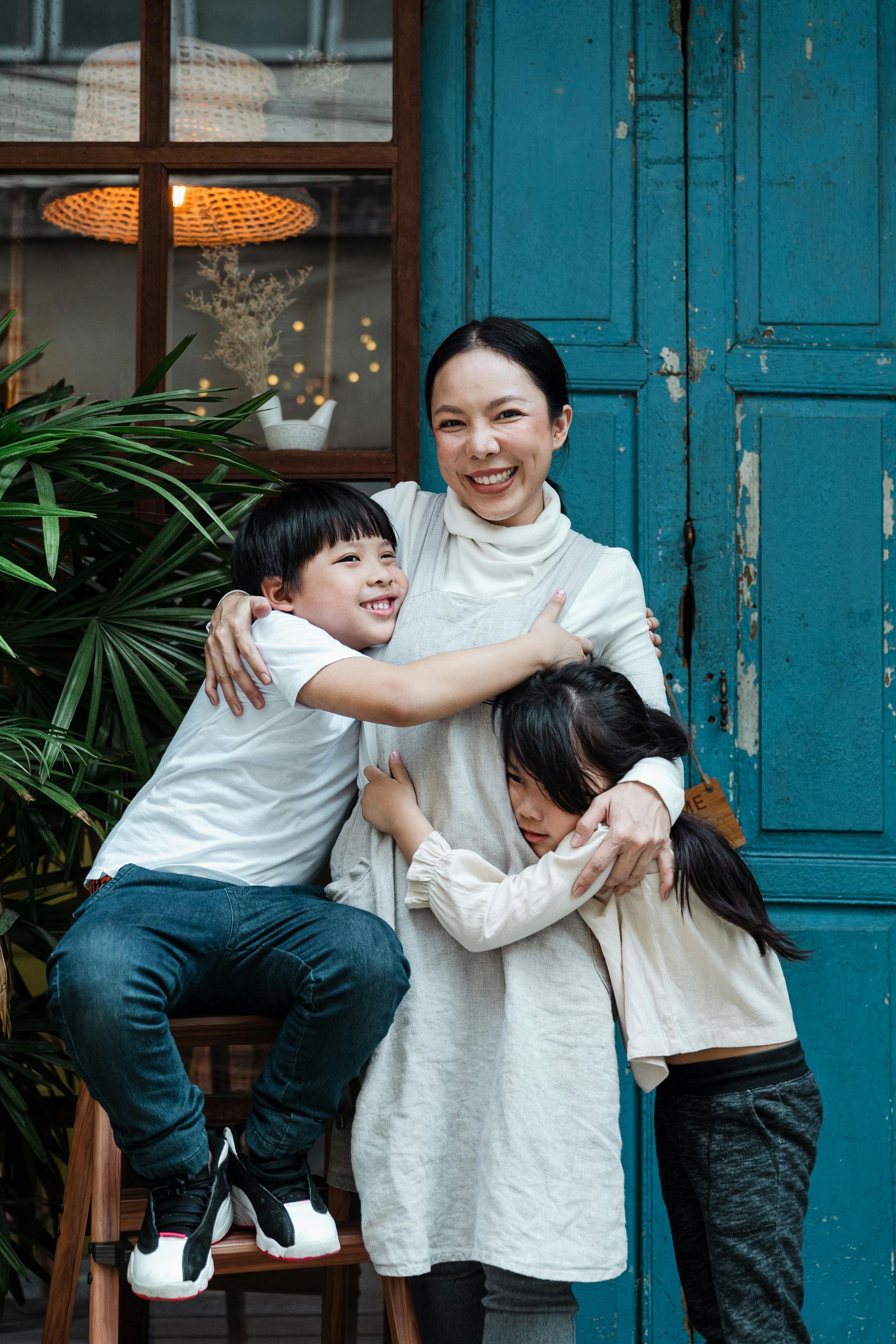 Woman with children hugging, smiling near blue door, greenery.