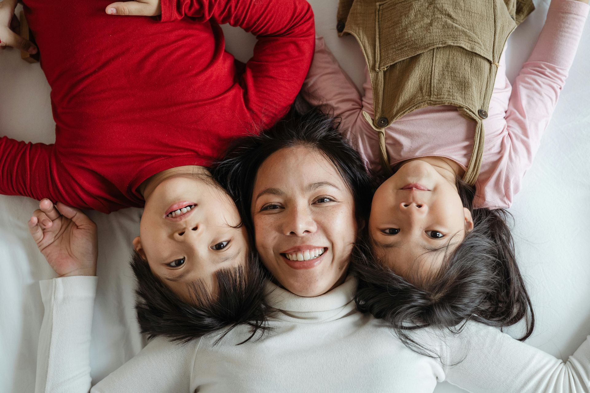 Mother and two children lying on a bed, smiling. Red shirt, green overalls.